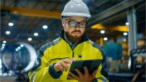 Experienced electrician in Perth using a tablet, wearing safety gear, inspecting electrical equipment in an industrial environment.